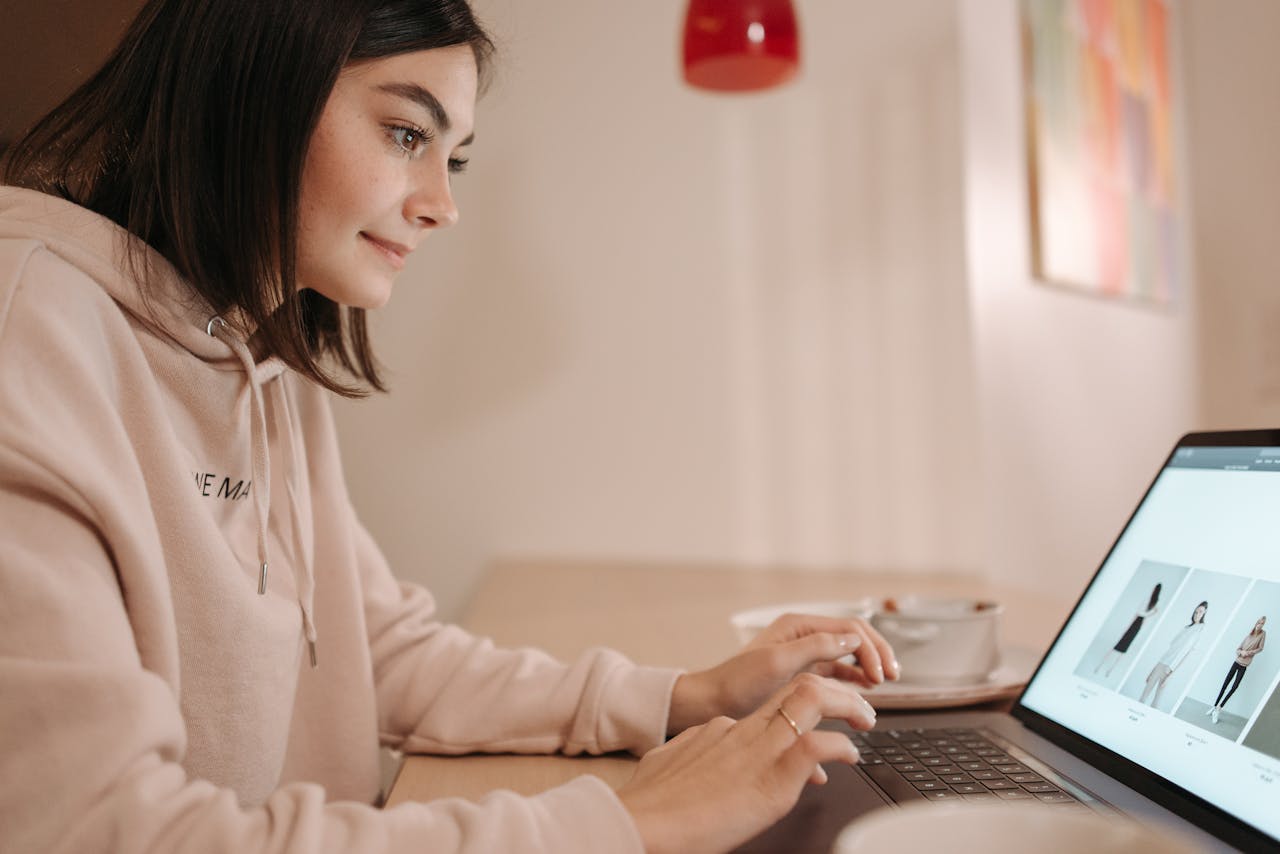 gallery-6 Young woman in pink hoodie browsing online fashion store on laptop at home. Calm and focused atmosphere.
